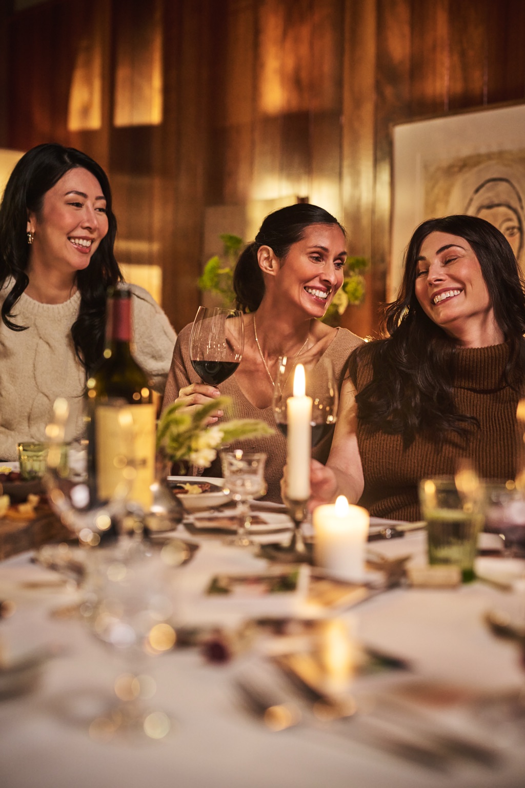 Three women sharing a laugh over glasses of Duckhorn cabernet at a candlelit dinner table, photographed by Dawn Heumann