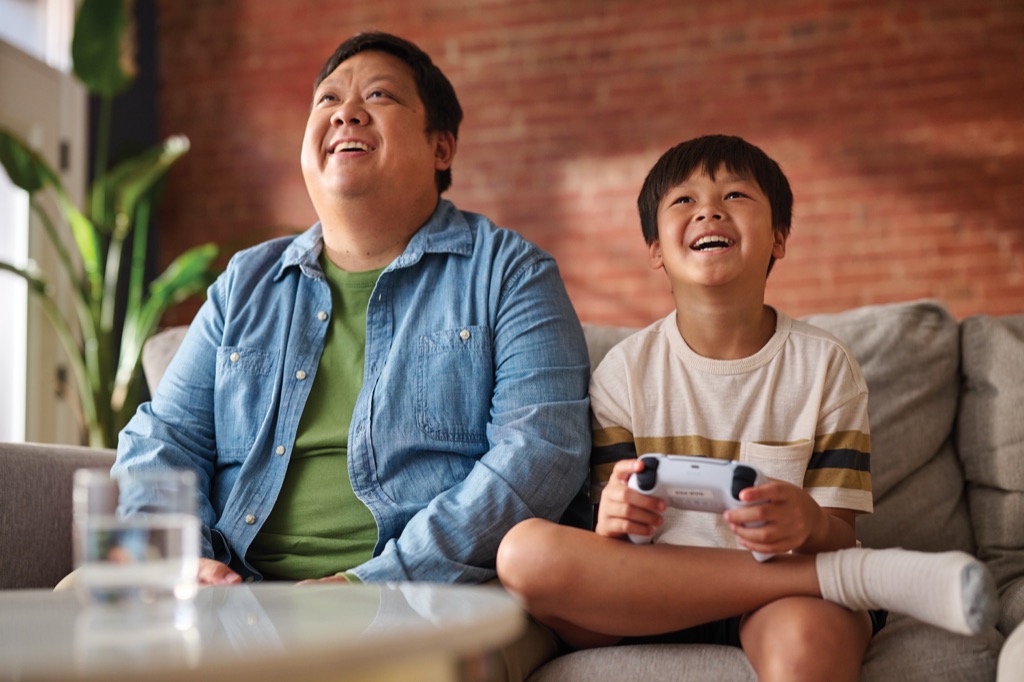 Father and son looking up laughing as the boy plays a video game from the couch, photographed by Dawn Heumann