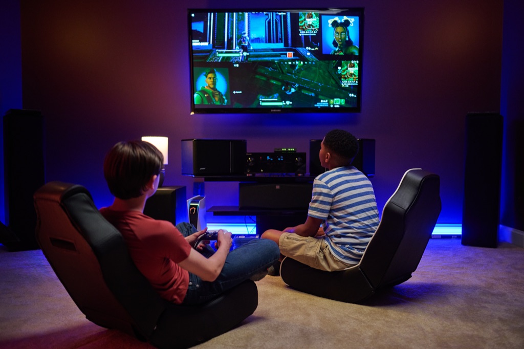 Two boys in floor rockers playing a co-op video game on a wall-mounted TV in a purple-lit den