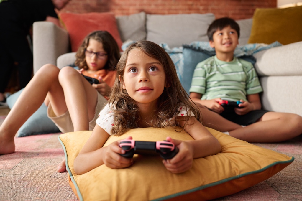 Young girl resting on a marigold floor pillow playing a video game with friends behind her