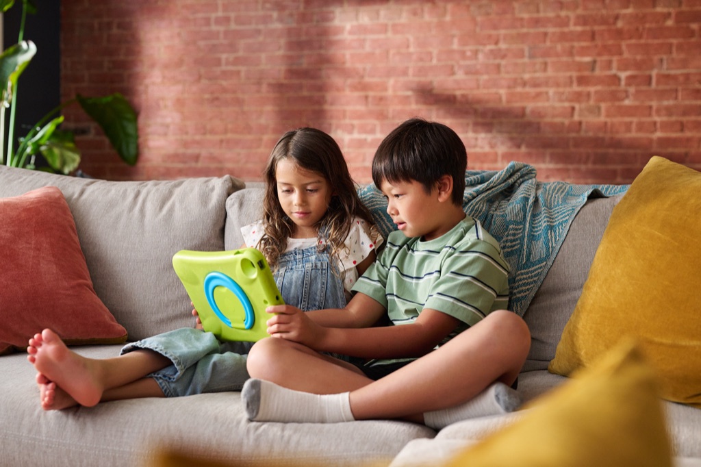Two kids sharing a tablet in a green kid-proof case on the living room sofa