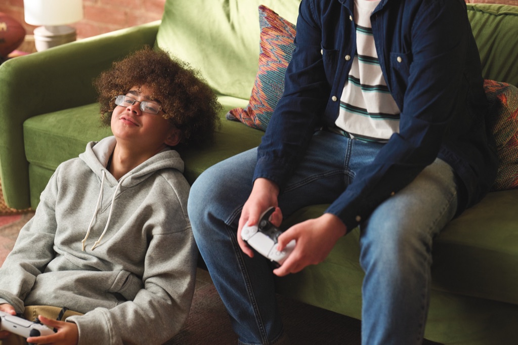 Teen grinning up from the floor with a controller while his friend plays from the green sofa