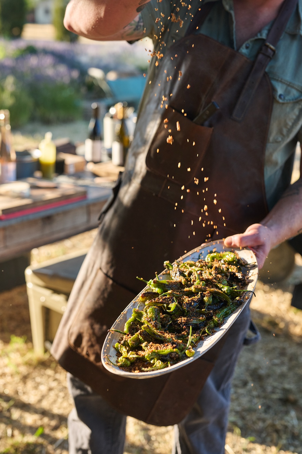 Chef sprinkling toasted breadcrumbs over a platter of blistered peppers beside a lavender field