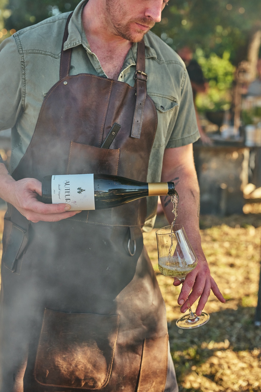 Chef pouring Auteur Chardonnay into a glass through drifting grill smoke at an outdoor dinner