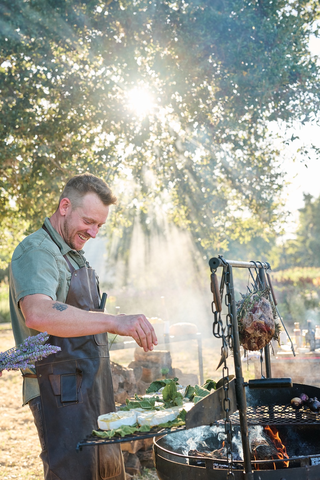 Smiling chef seasoning cheese on fig leaves over a swinging live-fire grill under a sunlit oak tree