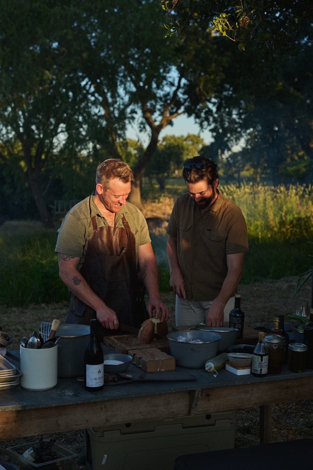 Two chefs laughing while slicing bread at an outdoor prep table in the last light of day