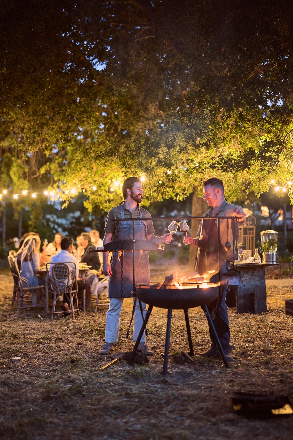 Two chefs toasting glasses of wine over the fire pit at night under string lights, photographed by Dawn Heumann