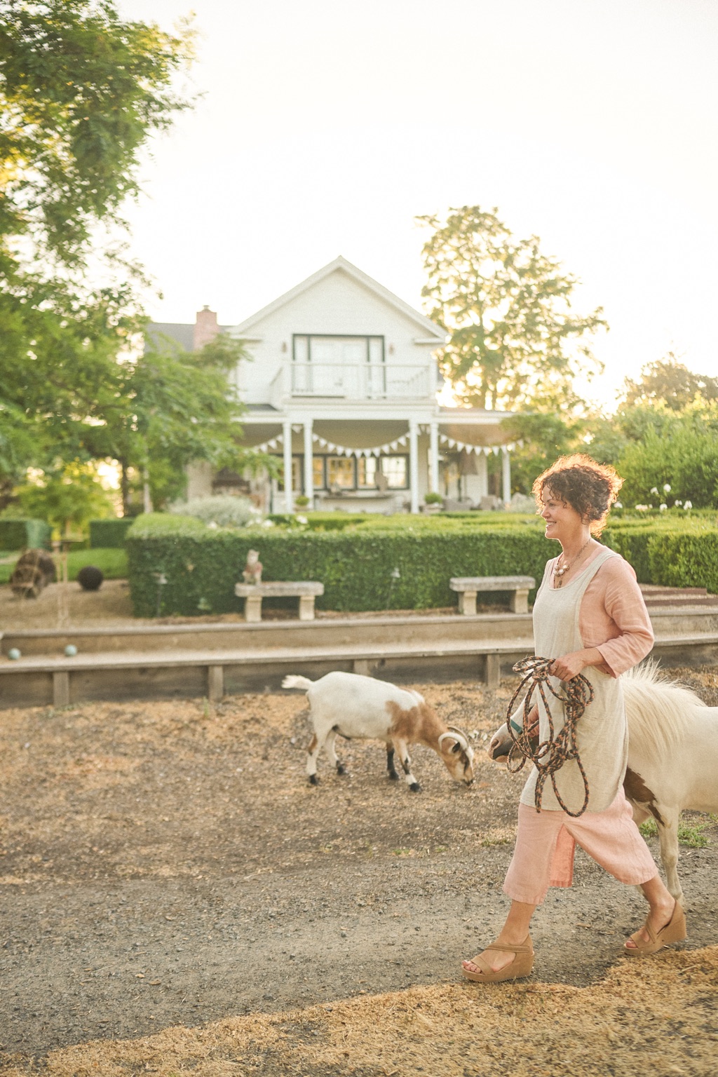 Woman leading a pony past a grazing goat in front of a white farmhouse strung with bunting