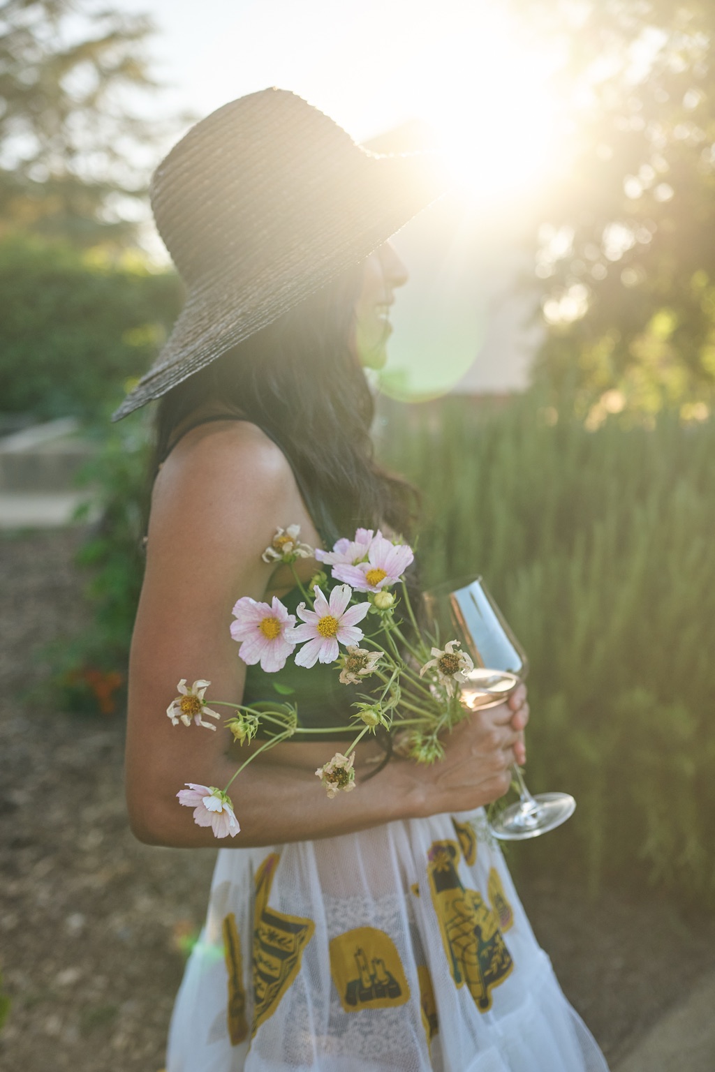 Woman in a wide-brim straw hat carrying cosmos flowers and a glass of rose through the garden at sunset