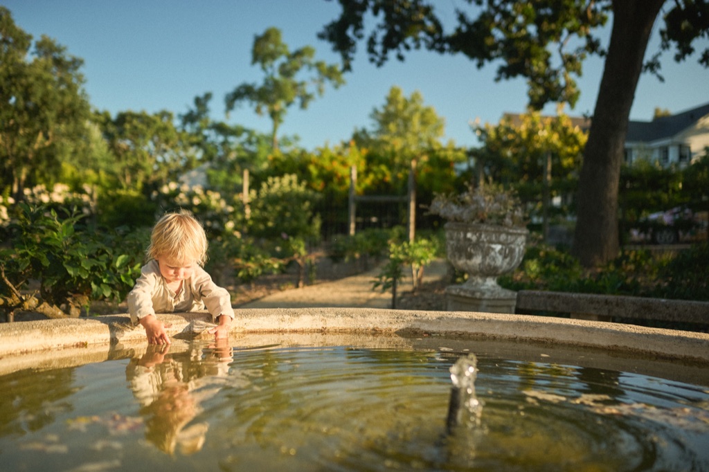 Toddler playing in a stone garden fountain at golden hour with vineyard rows behind