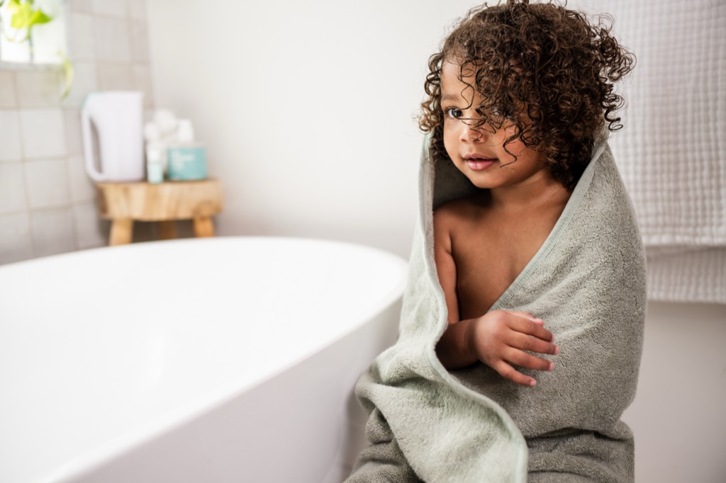 Toddler with curly hair wrapped in a sage hooded towel beside the bathtub, Frida Baby campaign