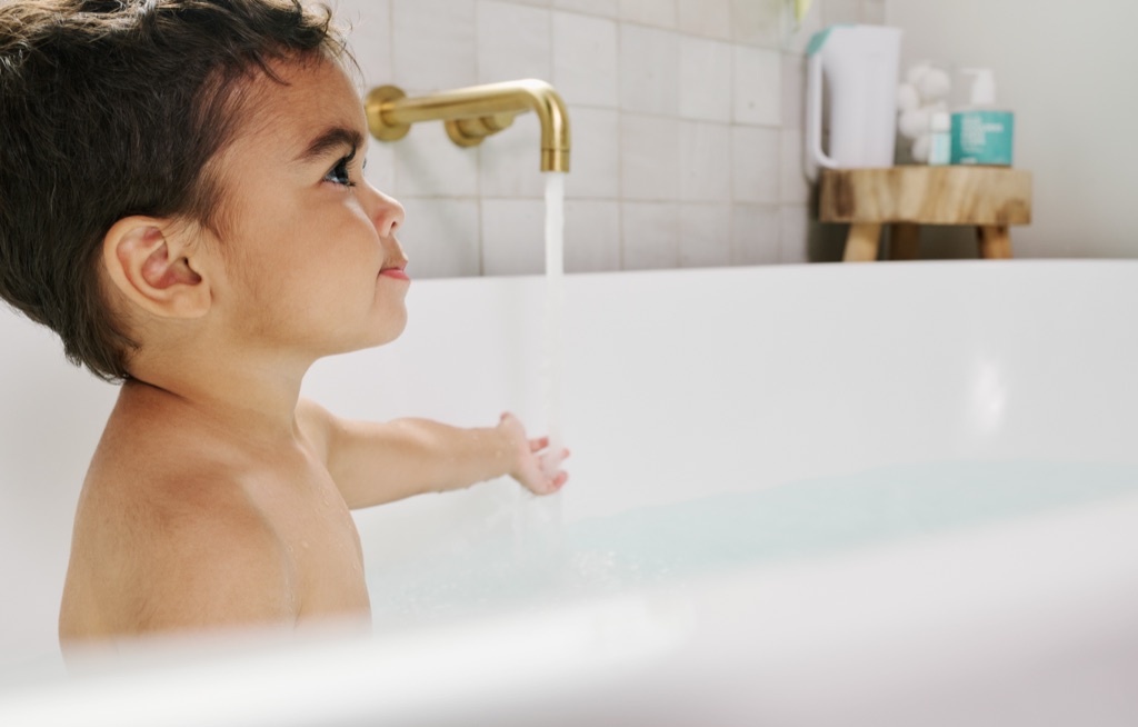 Toddler in the bathtub watching water pour from a brass faucet, Frida Baby bath campaign
