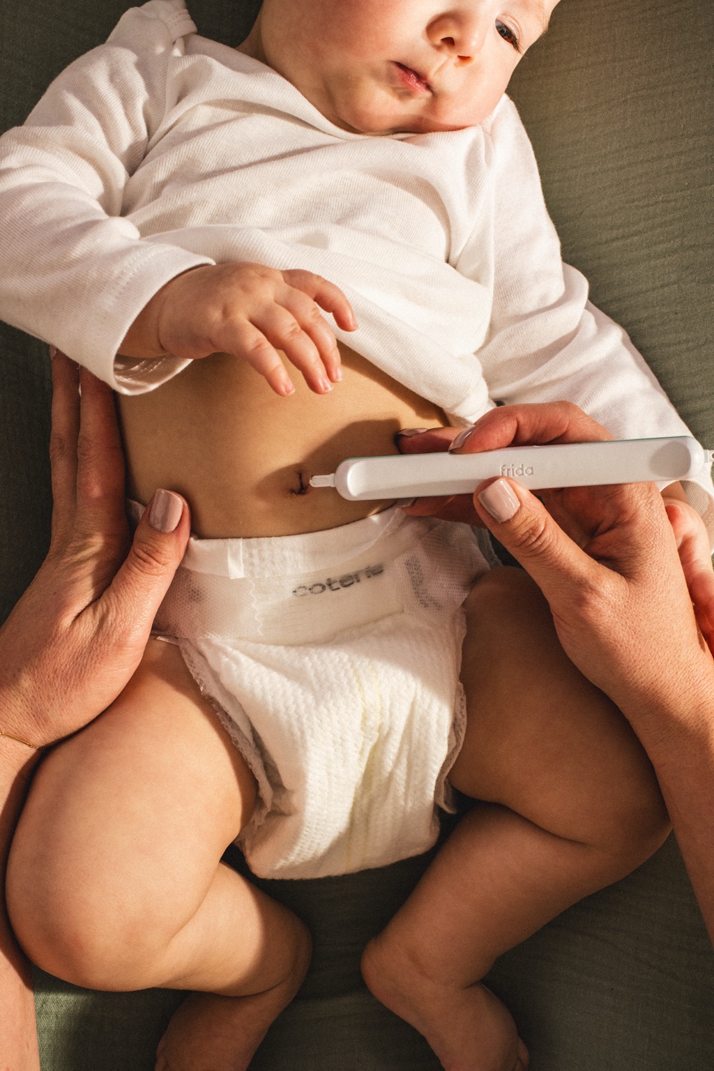 Overhead detail of a Frida Baby belly care device on a newborn's navel with an olive muslin blanket