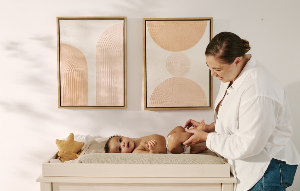 Mother tending to her baby at a changing table in a warm modern nursery, Frida Baby campaign