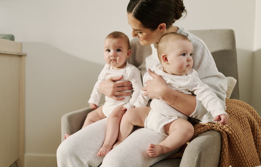 Mother on a glider holding twin babies in white onesies, Frida campaign photography