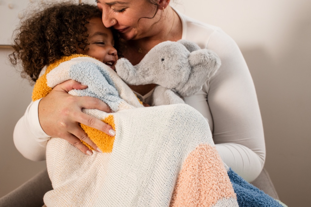 Mother hugging her laughing toddler wrapped in a knit blanket with a plush elephant, Frida campaign