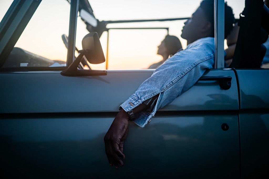 Man's arm in a denim jacket draped over the door of a sage green Bronco at dusk, friends silhouetted against the sunset