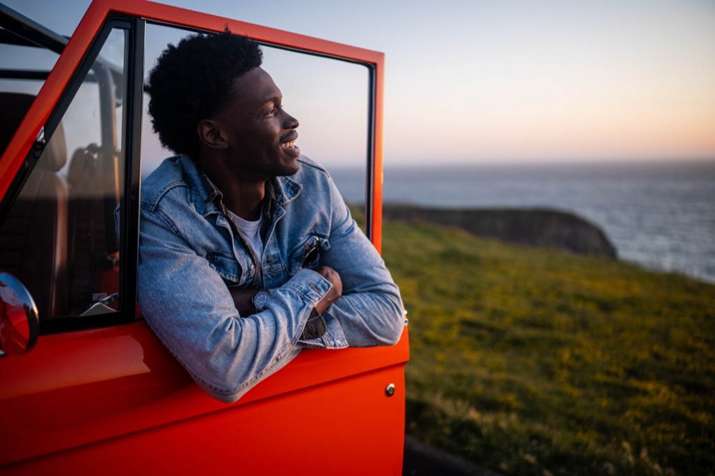 Man in a denim jacket leaning on the open door of an orange Bronco looking out at the Pacific at sunset