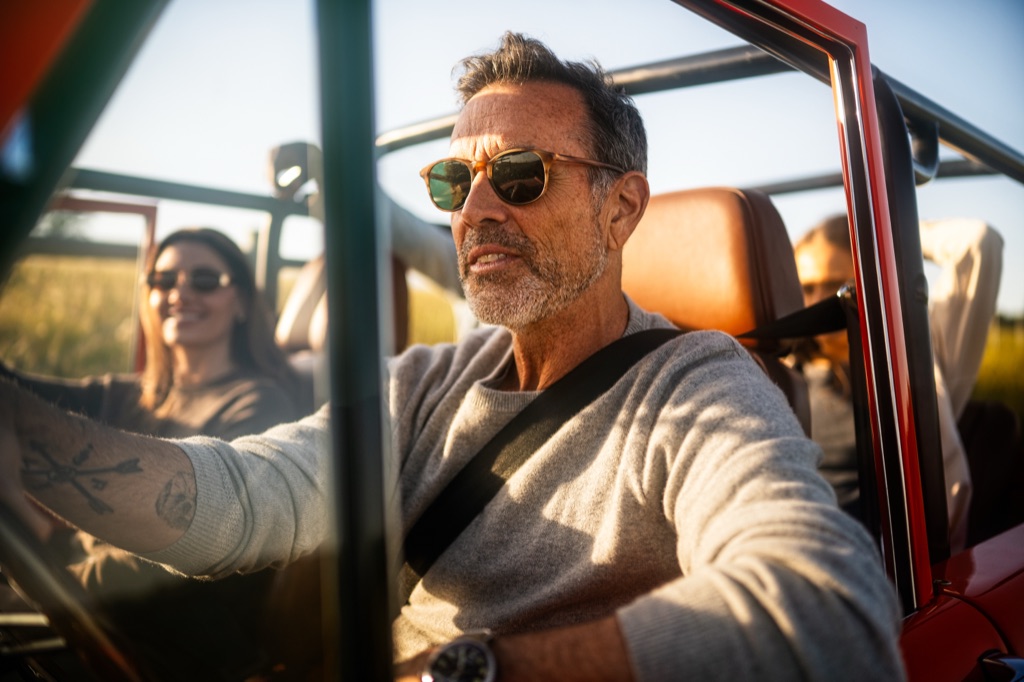 Silver-haired man in sunglasses driving a restored orange Ford Bronco with friends at golden hour