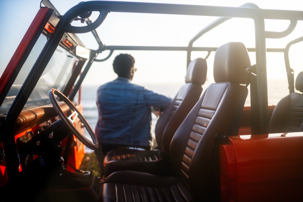View across the leather seats of an orange vintage Bronco to a man in a denim jacket gazing at the ocean