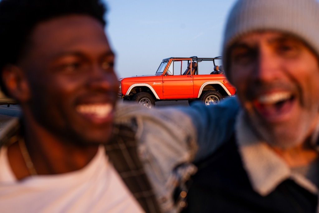 Two men laughing at dusk with an orange vintage Ford Bronco parked in profile behind them