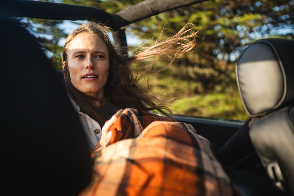 Woman with hair blowing in the wind riding in the back seat of an open-top vintage Bronco with a plaid blanket on her lap