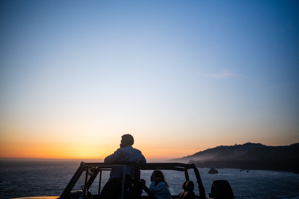 Silhouette of a man standing in the roll cage of an open-top Bronco against a dusk sky over the ocean, photographed by Dawn Heumann