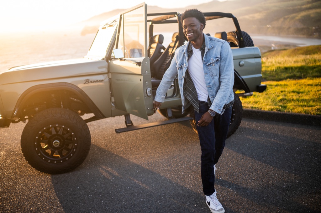 Man stepping out of a sage green vintage Ford Bronco on a coastal bluff at golden hour