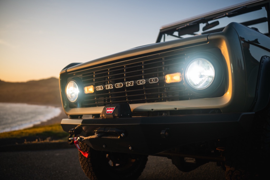 Front grille of a restored sage green Ford Bronco with headlights on and a winch at coastal dusk