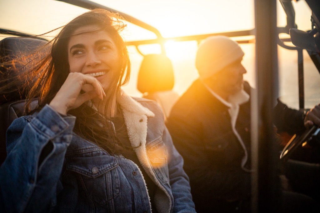 Woman in a sherpa collar denim jacket smiling in the back seat of a vintage Bronco at sunset, heavy sun flare