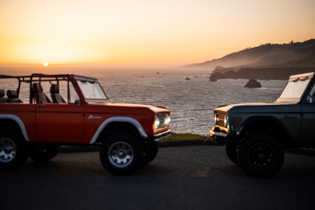 Orange and sage green vintage Ford Broncos parked nose to nose at a Sonoma Coast overlook as the sun sets over the Pacific