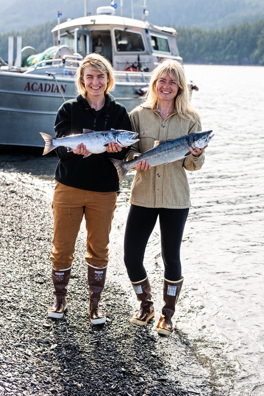 Two women on a pebble beach holding salmon in front of the beached fishing boat Acadian, both in XtraTuf boots on the Alaska coast