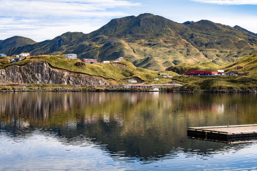 Green treeless Aleutian hills with homes above a rocky shoreline reflected in a calm harbor on the Alaska coast