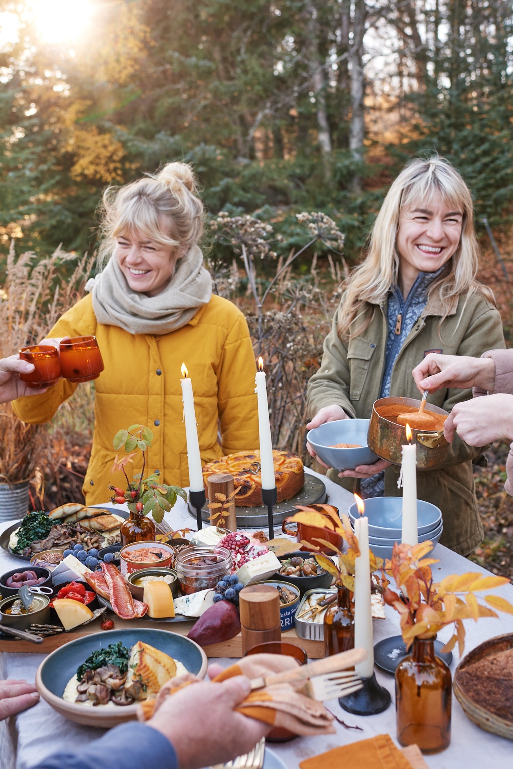 Two women laughing at a fall outdoor table as one raises amber glasses and the other serves soup from a copper pot