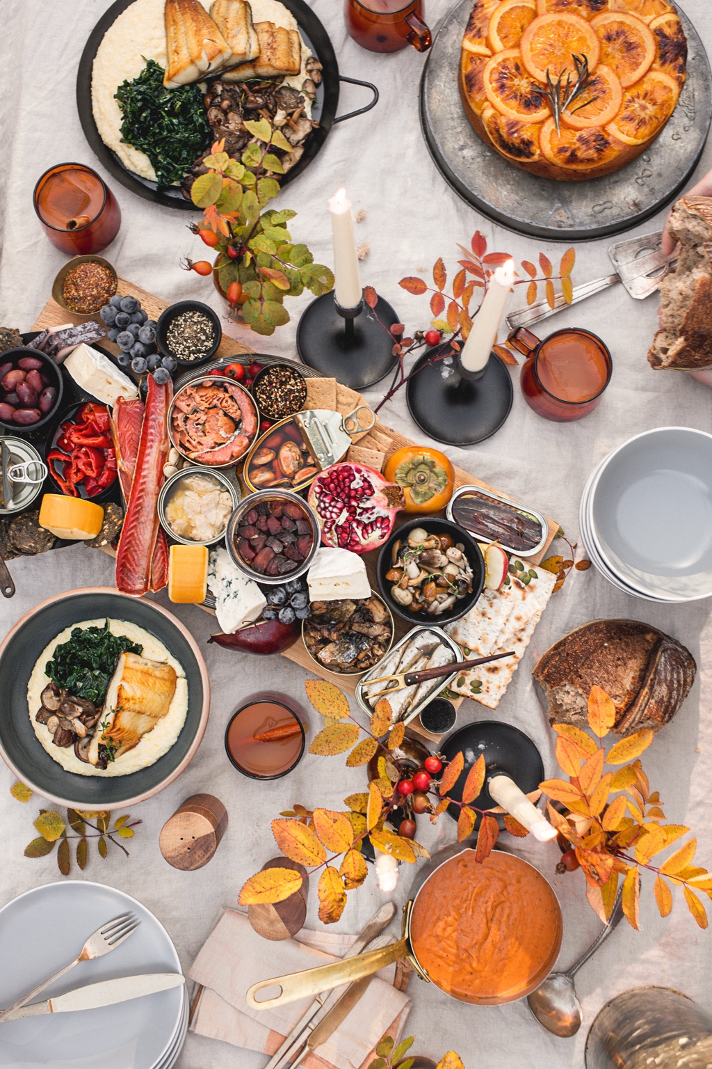 Overhead autumn feast with a tinned fish and smoked salmon board, black cod over polenta, orange upside-down cake, and taper candles