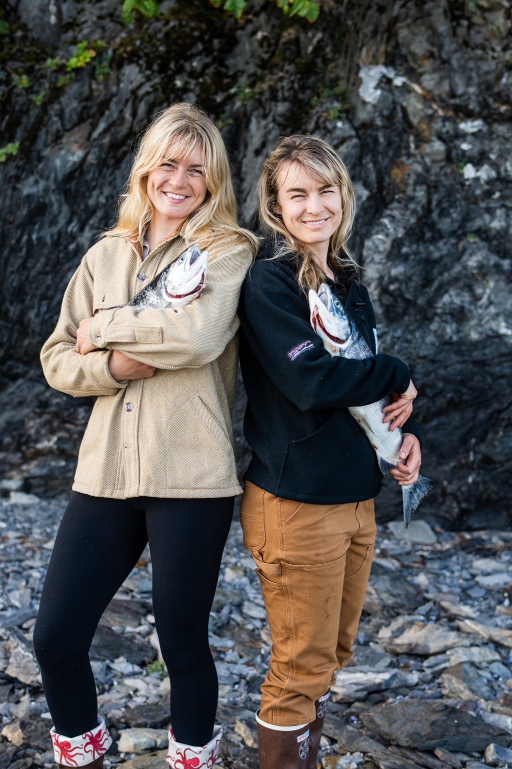 Two smiling women standing back to back against a rock face, each cradling a fresh salmon, photographed in Alaska