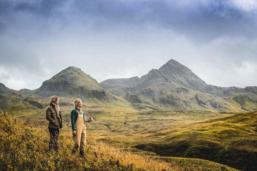 Two women walking golden tundra hills below rugged peaks, one carrying a berry pail