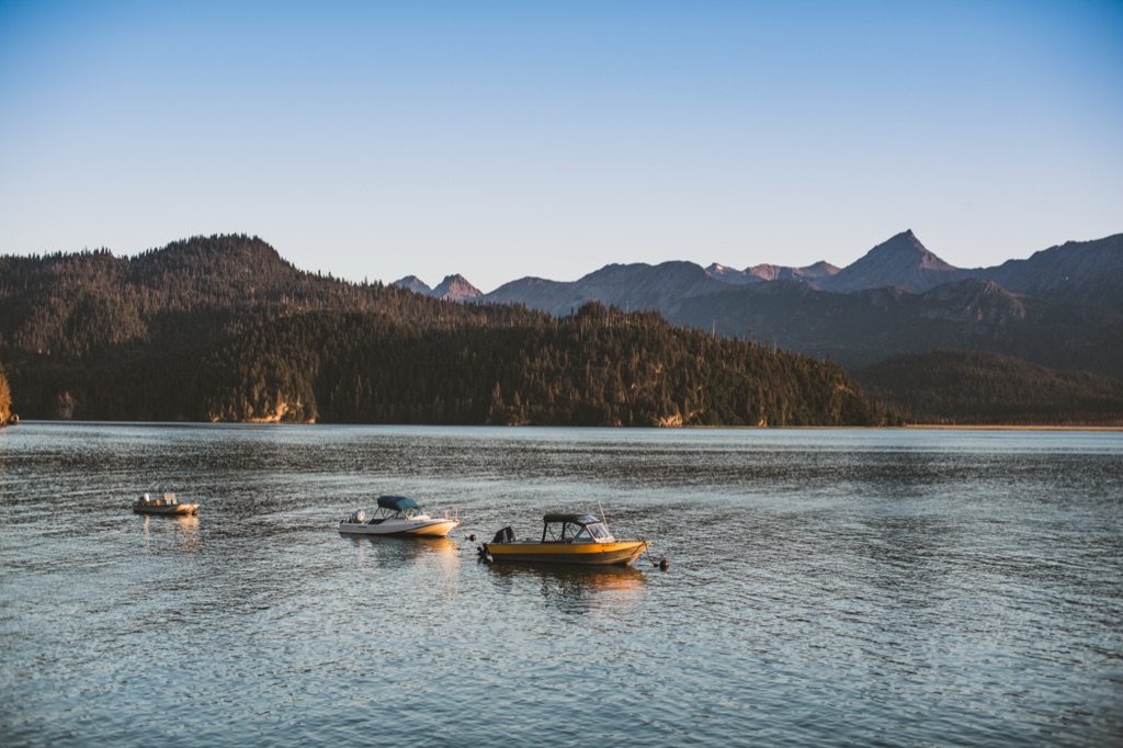 Three small boats moored on a calm bay at dusk beneath forested hills and peaks in Alaska