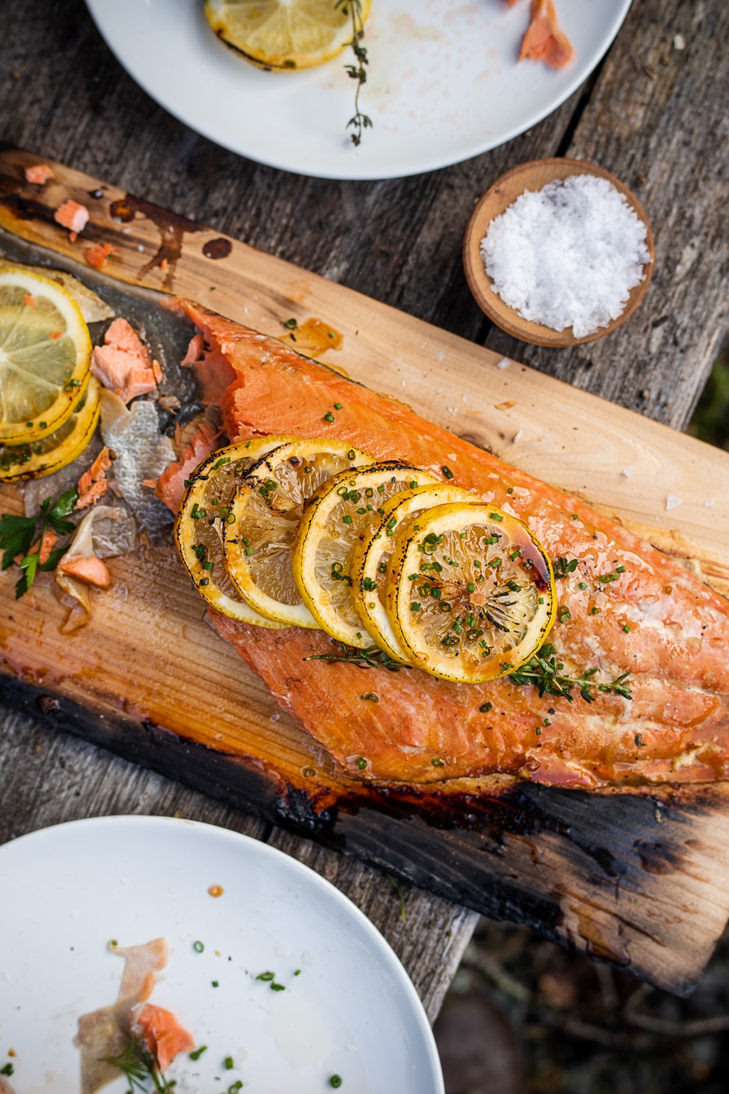 Cedar plank salmon topped with charred lemon slices, chives, and thyme on a weathered picnic table, photographed in Alaska
