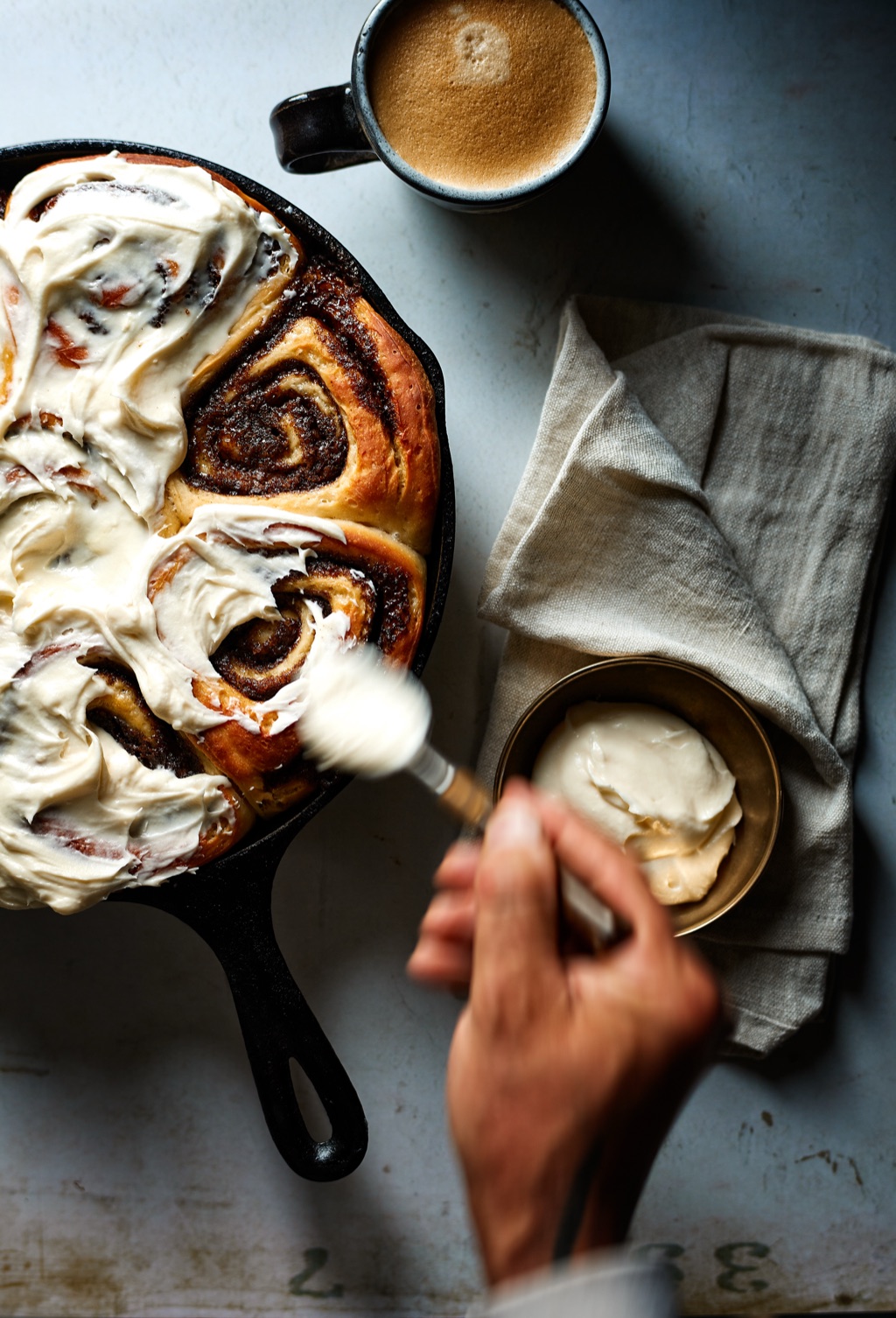 Brushing cream cheese frosting over cinnamon rolls in a cast iron skillet beside a mug of coffee
