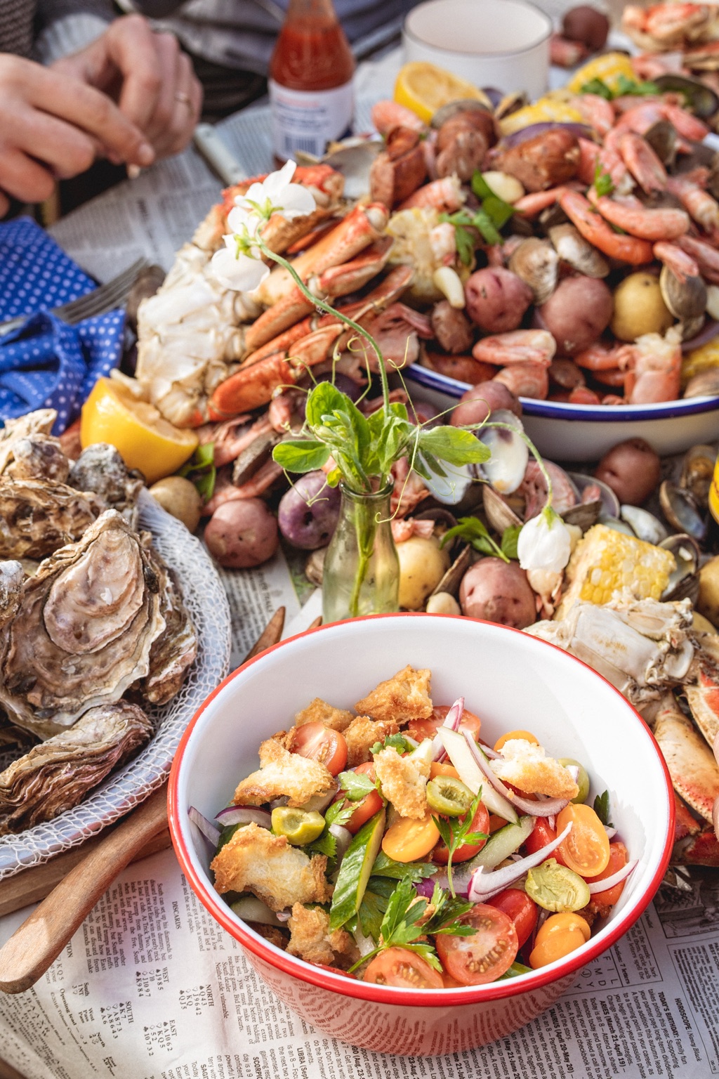 Seafood boil spread with crab legs, shrimp, potatoes, and a bowl of tomato panzanella beside oysters and a pea shoot posy