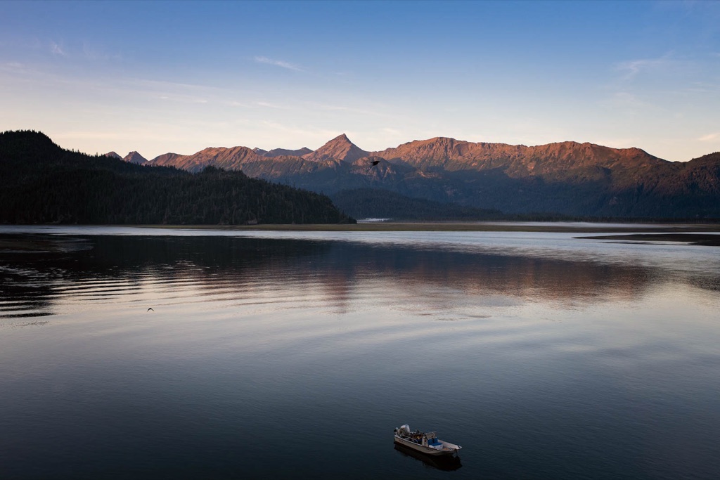Mirror-calm bay at dawn with alpenglow on the mountain range and a lone skiff at anchor in Alaska