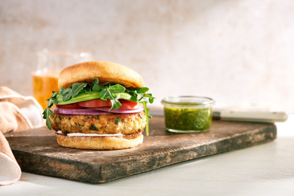 Herbed fish cake burger with arugula, avocado, tomato, and red onion on a brioche bun beside a jar of salsa verde
