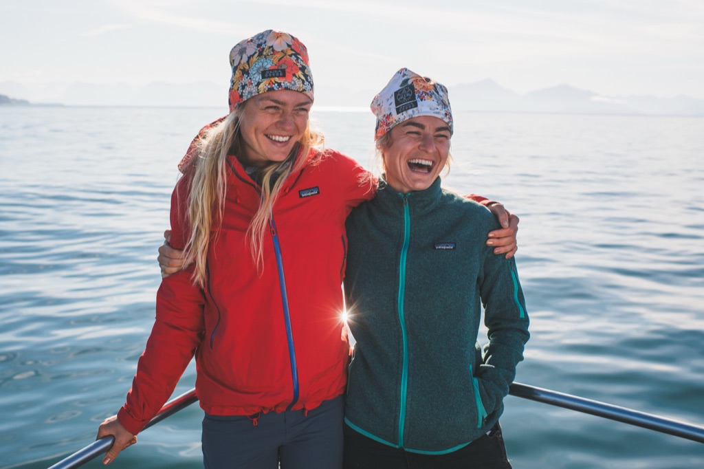 Two women laughing arm in arm on a boat bow in floral beanies and Patagonia jackets in Alaska