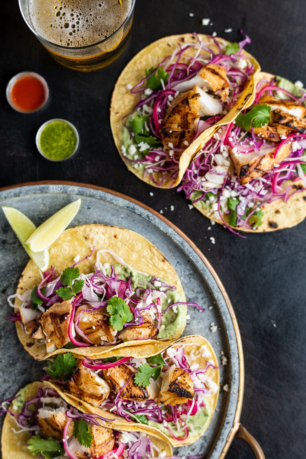 Grilled fish tacos on corn tortillas with red cabbage slaw, pickled onion, cotija, and cilantro beside a glass of beer