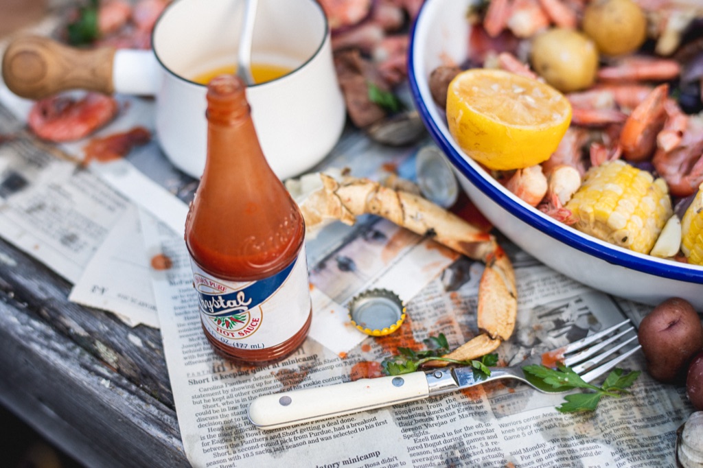 Crystal hot sauce bottle and a picked crab claw on newspaper beside a pot of drawn butter and a bowl of shrimp and corn in Alaska