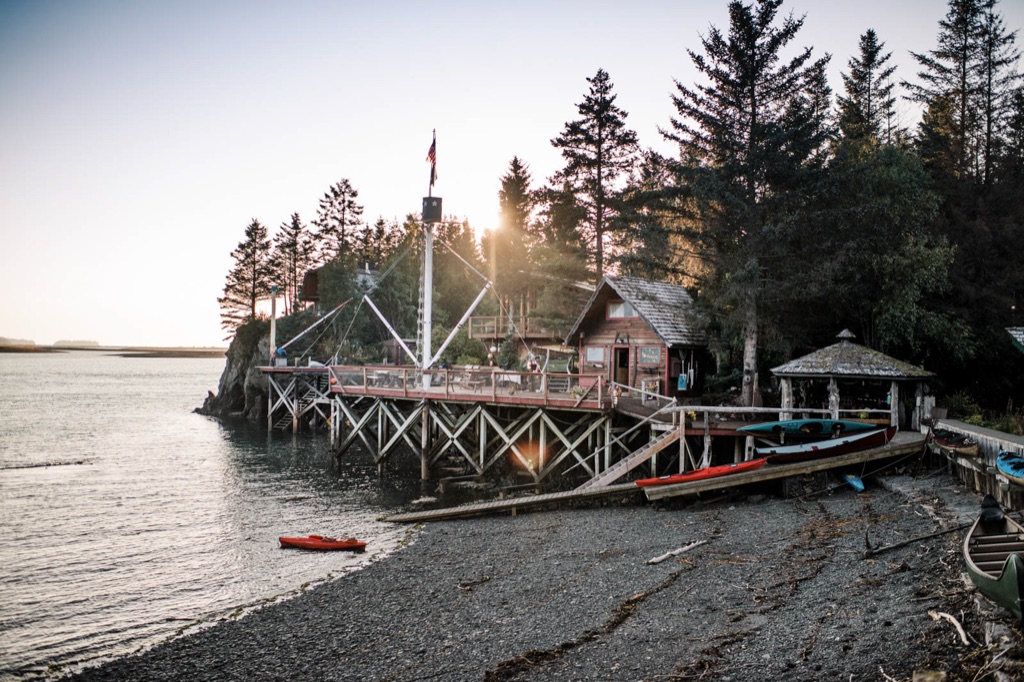 Waterfront kayak lodge on wooden pilings at sunset in Kachemak Bay, Alaska, with kayaks lined up on the gravel beach