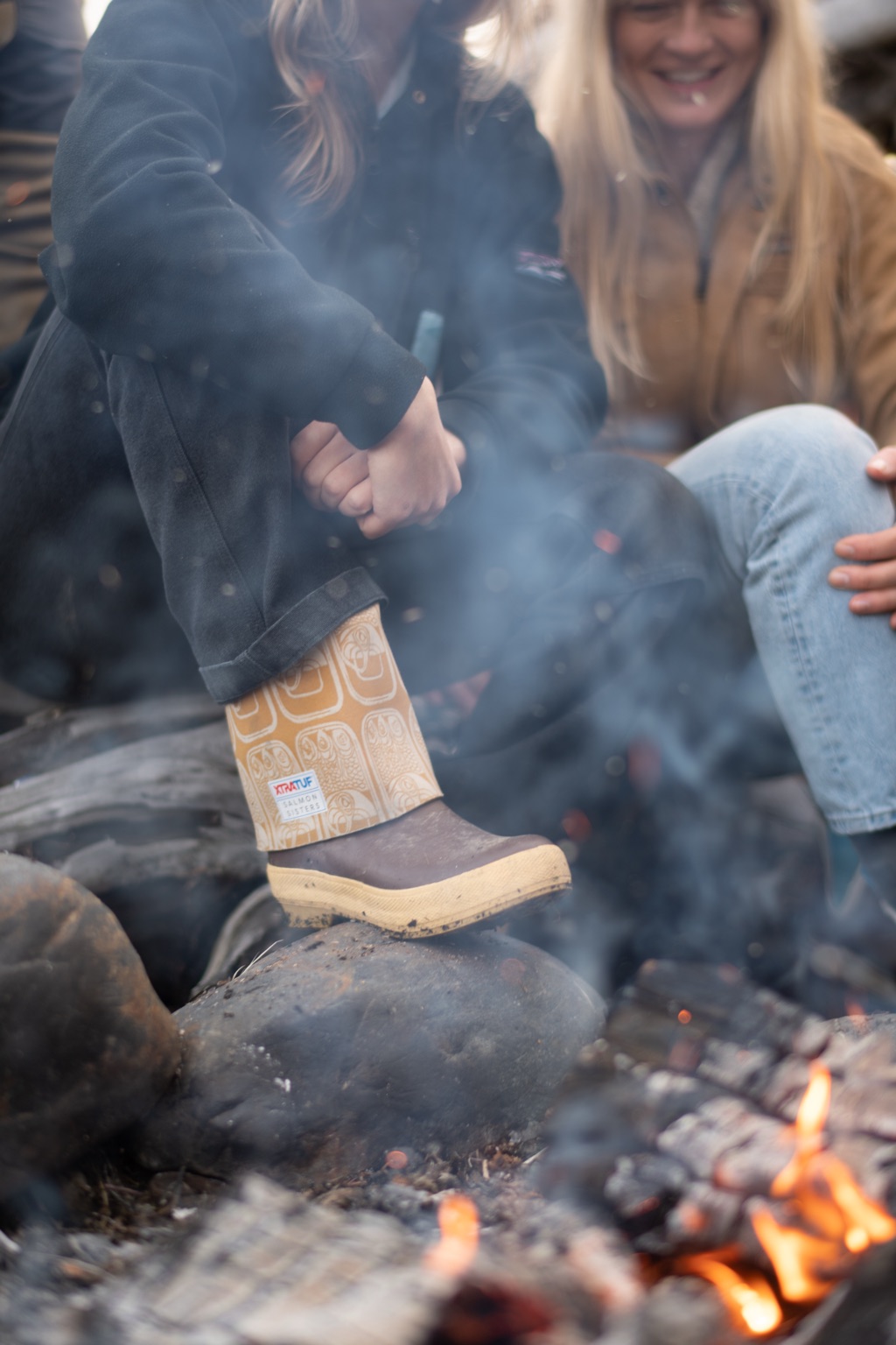 Child's XtraTuf Salmon Sisters boot with a tin can print cuff resting on a rock beside a smoky campfire