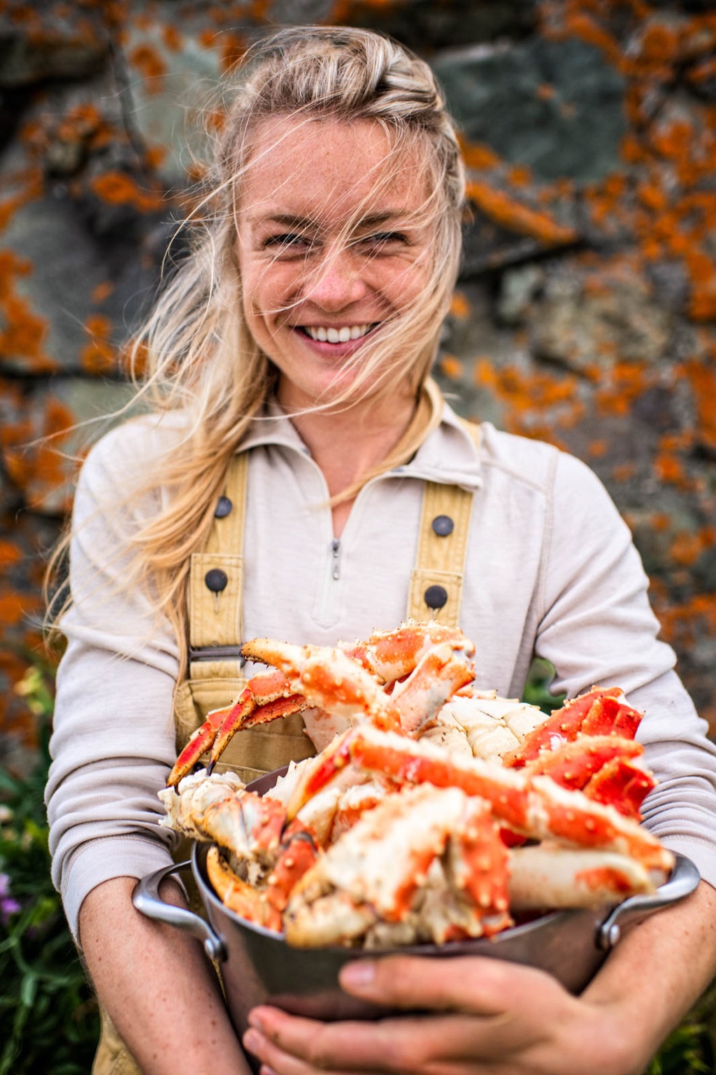 Woman holding a colander piled with king crab legs against lichen-covered rock