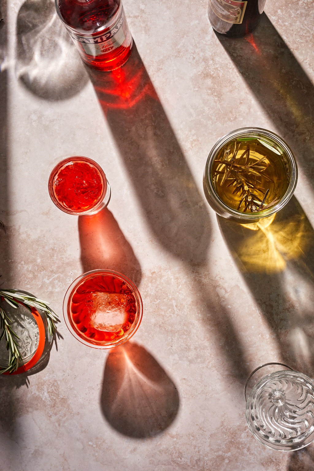 Overhead of negronis and a rosemary infusion in hard sunlight with long shadows and a Campari bottle